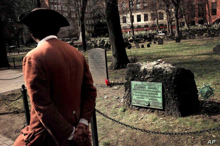 Samuel Ike, of Cambridge, Mass., left, dressed in the role of Revolutionary War-era African American abolitionist Prince Hall, walks past the grave with red flowers of the victims of the 1770 Boston Massacre, March 3, 2020.